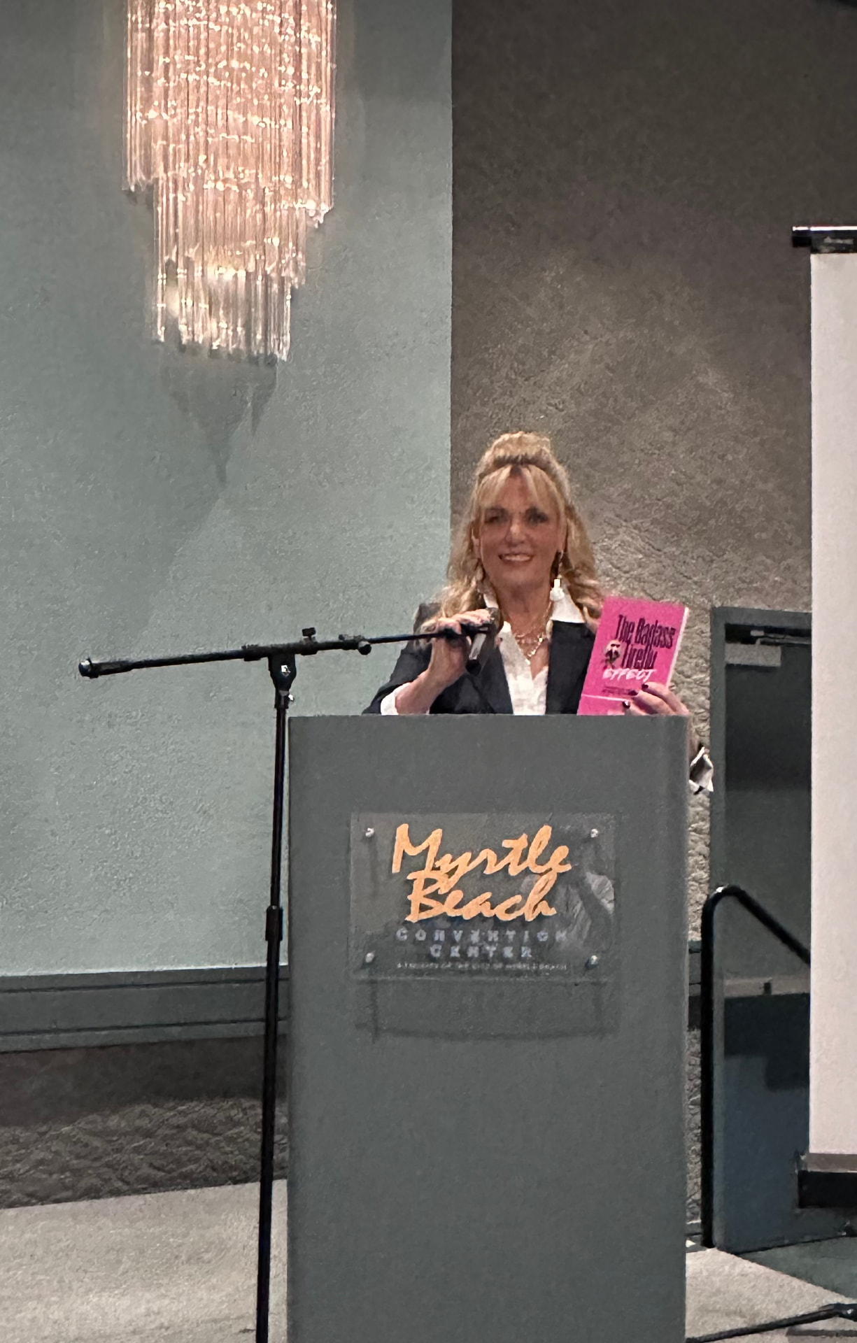 Jerri at the Myrtle Beach Convention Center podium, holding up a hot-pink copy of The Badass Firefly Effect.