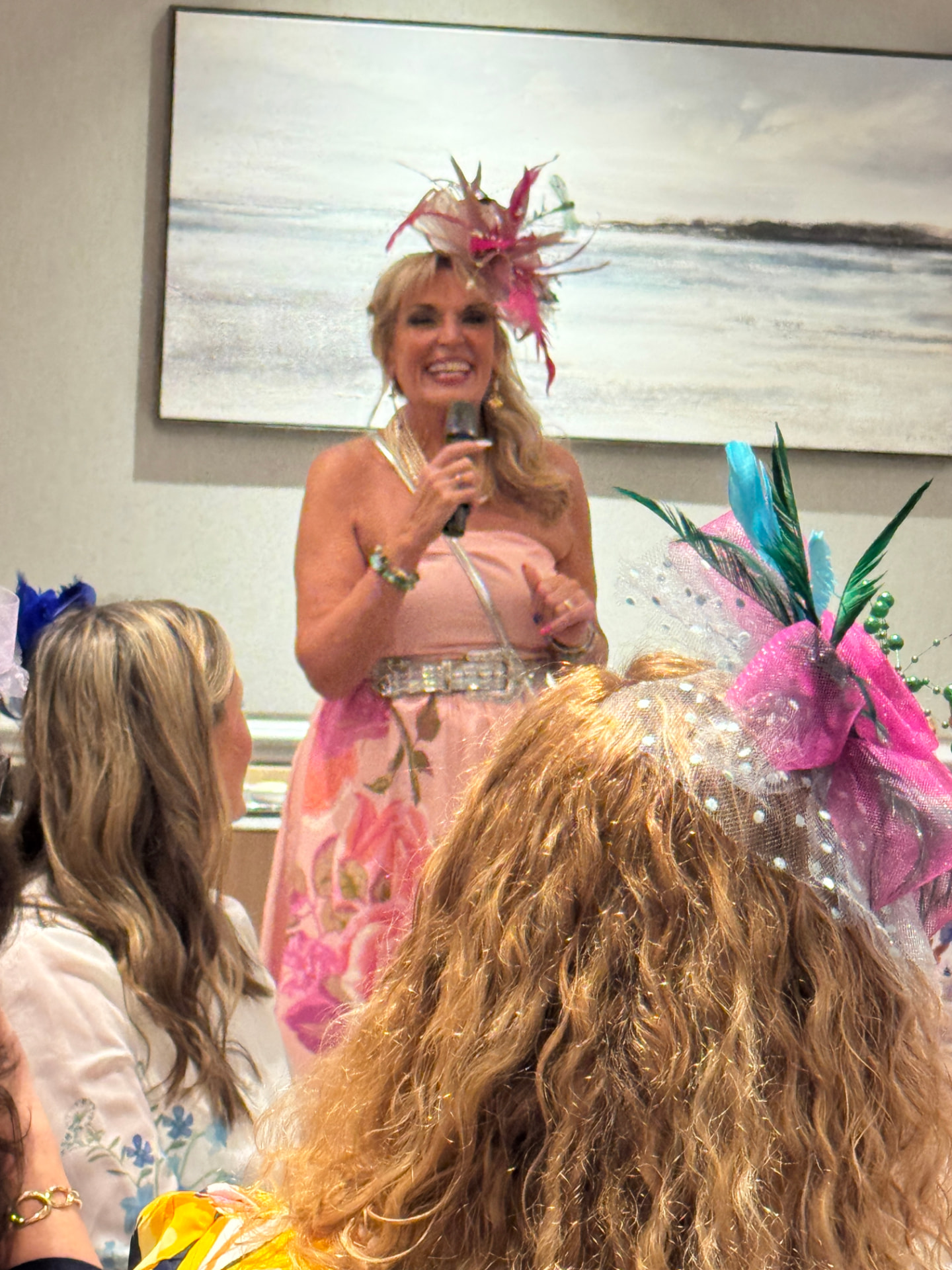 Jerri speaking into a mic in a floral dress and feathered fascinator hat, with participants in Derby hats in the foreground — a joyful, colorful moment.