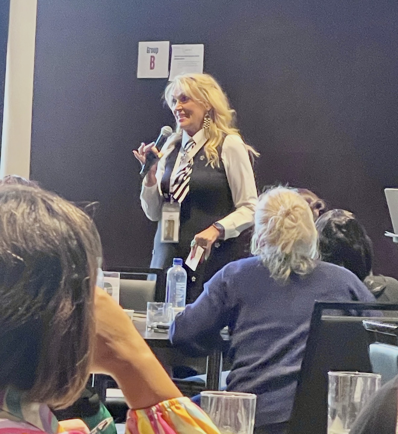 Jerri standing among round tables of participants, microphone in hand, mid-conversation with the room.