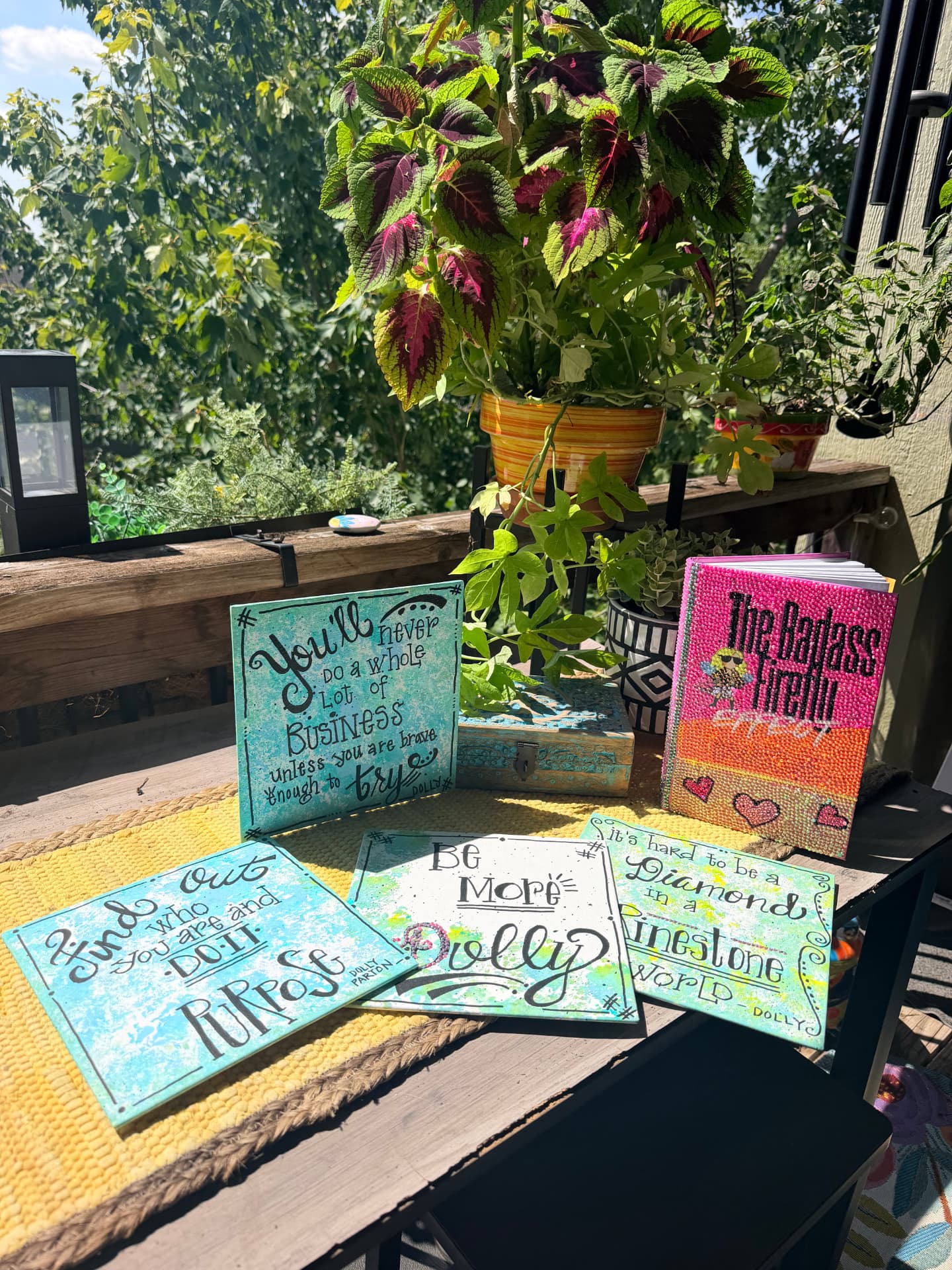 Five handmade, hand-lettered Dolly Parton quote canvases displayed on a sunny deck table beside The Badass Firefly Effect book