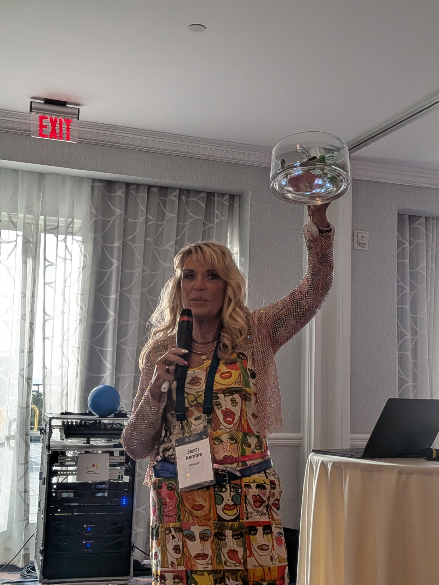Jerri holding a real glass firefly jar high above her head mid-keynote, in a colorful artist-print dress.